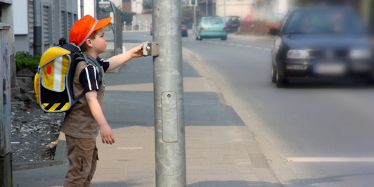 Auf dem Bild ist ein Schuljunge zusehen, der an einer Ampel steht und die Straße überqueren möchte.